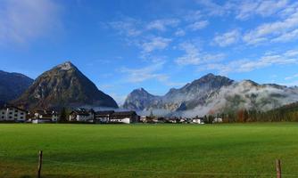 A picturesque landscape with green meadows and high mountains. The sky is clear and blue, and mist hovers over the valleys.