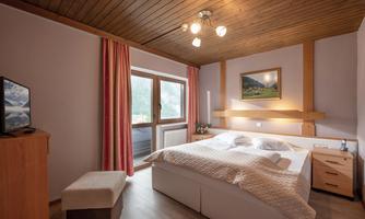 A cozy bedroom with a large bed and wooden floor. The room has windows with curtains and a warm, inviting atmosphere.