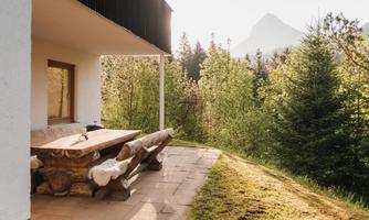 A cozy terrace with a wooden table and benches, surrounded by trees and mountains. The sky is bright and the atmosphere feels peaceful.