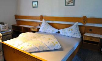 A simple bedroom with a large bed and two pillows. Small pictures hang on the wall, and there is a dresser and a table.