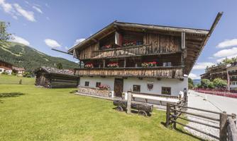 A traditional wooden house in the Alps with balconies and flowers. In the background, green hills and a blue sky can be seen.