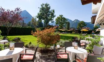An inviting garden with tables and chairs, surrounded by trees and mountains. The sun is shining brightly in the blue sky.