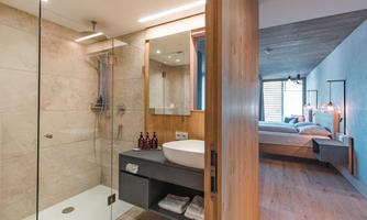 A modern bathroom with a glass shower and an elegant sink. In the background, there is a bedroom with wooden details.