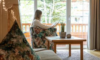 A woman is sitting in a cozy armchair and reading a book. The view looks out onto a balcony with green plants.