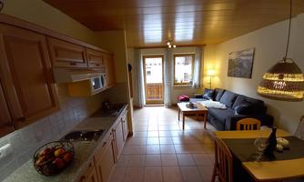 A cozy living kitchen with bright wooden cabinets and a dining area. In the background, there is a sofa and a balcony with a view outside.