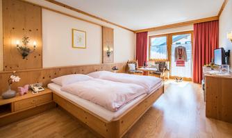 A cozy bedroom with a double bed and wood paneling. The room has large windows with access to a balcony and is bright and inviting.