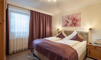 A cozy bedroom with a large bed and purple bedding. The room has bright curtains and a decorative wall with a picture.