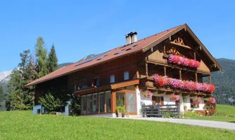 A traditional wooden house with beautiful balconies and colorful flowers. The surroundings are green and surrounded by mountains, under a radiant blue sky.