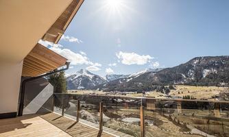 A view of the mountains with bright sunshine and a blue sky. In the foreground, there is a balcony with a glass railing.