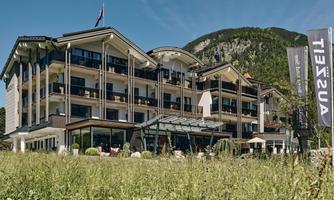 A modern hotel building surrounded by green meadows and mountains. The balconies offer a view of the natural surroundings.