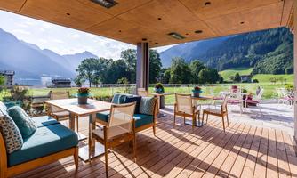 A modern outdoor area with a wooden terrace and comfortable seating. In the background, impressive mountains and a green landscape can be seen.