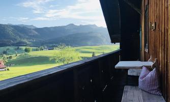 A cozy balcony with wood paneling overlooks green meadows and gentle hills. In the background, mountains and a blue sky are visible.