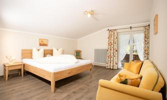 A bright bedroom with a large bed and a cozy sofa. The windows are adorned with curtains and provide natural lighting.