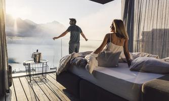 A couple enjoys the view of a picturesque lake from their modern bedroom. The atmosphere is tranquil and relaxing, with gentle rays of light shining through the windows.