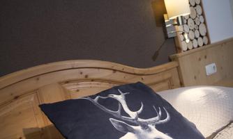 A cozy bedroom with a wooden bed and a decorative pillow featuring a deer motif. On the wall, a lamp and wood logs are visible.