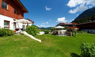 A picturesque garden area with a red wooden house and a green lawn. In the background, the mountains and a modern building can be seen.
