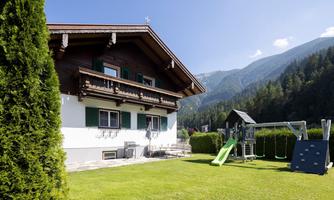 A beautiful chalet surrounded by green mountains. In the garden, there is a playground with a slide and a swing.