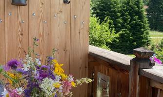 A cozy balcony with a wooden table and colorful flowers in a vase. In the background, mountains and a green forest landscape can be seen.