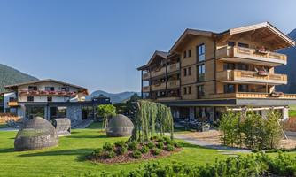 A modern wooden building with several floors surrounded by green gardens. In the background, gentle hills and a clear sky can be seen.