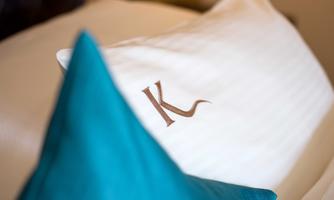 An elegant pillow with an embroidered "K" and a blue cushion in the background. The white bedding conveys a sense of comfort and style.
