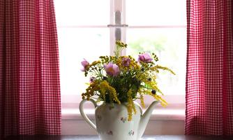 A pretty vase with colorful flowers stands on a table in front of a window. Red curtains frame the room and create a cozy atmosphere.