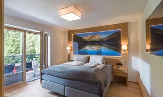 A modern bedroom with a large bed and a beautiful landscape picture on the wall. The room has large windows that lead to a balcony with a view of the greenery.