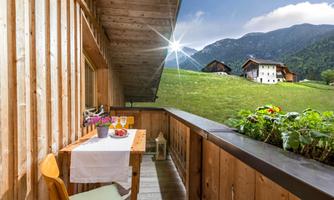 A cozy balcony with wooden-paneled walls and a view of green meadows and mountains. Drinks and fresh fruits are on the table, inviting for a relaxing time outdoors.
