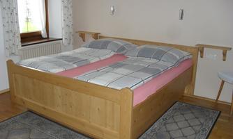 A bedroom with a large, bright wooden double bed. The room is simply and cozily furnished with carpets and windows that let in daylight.