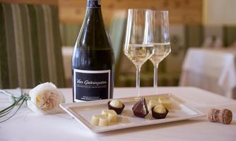 A bottle of sparkling wine is on a table next to two glasses. Various chocolates are arranged on a plate, accompanied by a rose.