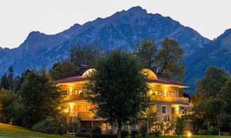 A charming house surrounded by trees, illuminated in the twilight. Majestic mountains rise in the background.