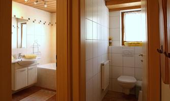 A bright bathroom with white tiles and a window. On the left side, a bathtub and a sink can be seen.