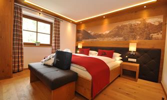 A stylishly furnished bedroom with wooden floors and modern furniture. The walls are decorated with a picture of mountains, and there are large windows with curtains.