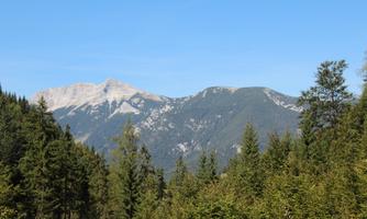 A picturesque mountain landscape with high peaks and dense forests. The clear blue sky contributes to the idyllic atmosphere.