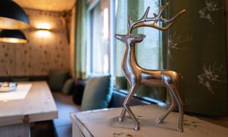 A stylish decoration in the shape of a silver deer stands on a table. In the background, there are windows with green curtains and a cozy seating area.