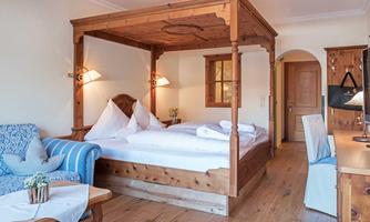 A cozy bedroom with a large wooden four-poster bed and white bedding. The room is bright and inviting, with a small sofa and warm wooden flooring.