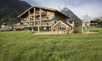 A modern building with balconies amidst a green meadow. In the foreground, there is a playground with wooden structures and a slide.