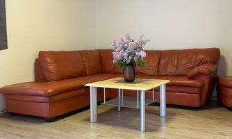 A cozy living room with a large brown leather sofa and a small wooden table. On the table stands a vase with colorful flowers.