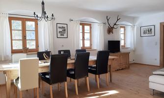 A bright dining room with a large wooden table and black chairs. Pictures hang on the walls, and a mounted antler is decorated.