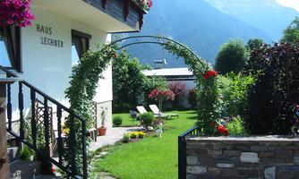 A pretty house with a well-maintained garden and a flower arbor. In the background, mountains and a clear sky are visible.