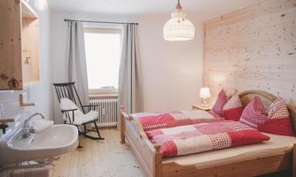 A cozy bedroom with a double bed and bright wooden paneling. There is an armchair and a small washing area.