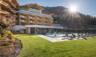 A modern hotel with a pool and loungers in the garden. The sun is shining over the mountain landscape.