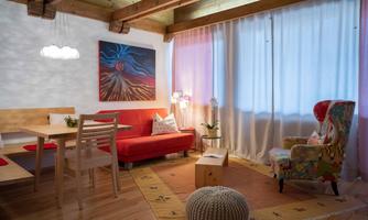 A cozy living room with a red sofa and a colorful armchair. Bright curtains let natural light in, and a piece of artwork adorns the wall.