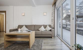 Bright, modern apartment with a cozy sofa and a wooden table. Large window doors offer a view of a snowy landscape.