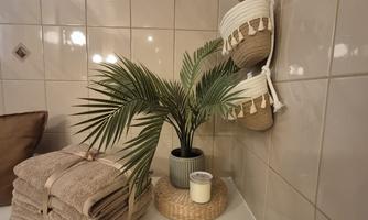A stylish household corner with folded towels and a small palm in a pot. In the background, decorative baskets and a candle holder are visible.