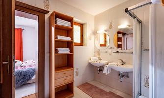 A modern bathroom with two sinks and large mirrors. In the background, a bedroom with a cozy bed can be seen.