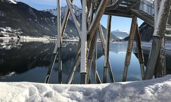 A snow-covered shoreline with a wooden walkway. In the background, the landscape is reflected in the calm water.