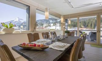 An elegant dining area with a large table and modern chairs. Large windows offer a beautiful view of the mountains and nature.