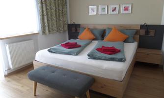 A modern bedroom with a double bed and colorful cushions. The walls are painted in neutral colors and there are wood and fabric elements for a cozy atmosphere.