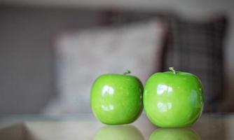 Two green candles shaped like apples are standing on a table. Blurry pillows are visible in the background.