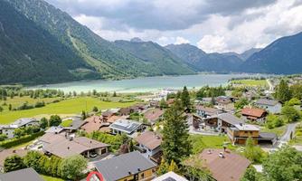 A picturesque view of a village in the mountains, surrounded by green meadows and a clear lake. In the background, majestic mountains rise under a cloudy sky.